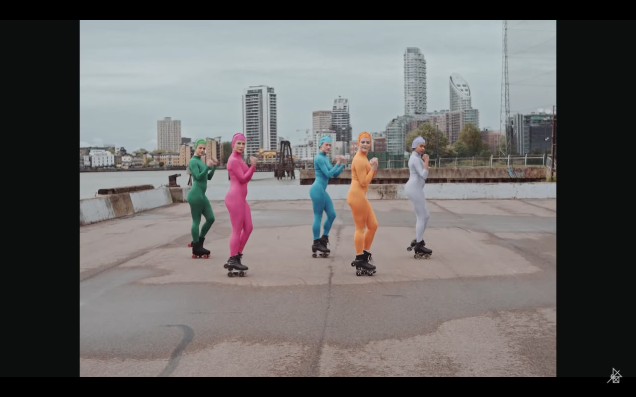 Still shot of four women wearing roller skates, each wearing different colored full body suits. City landscape in background