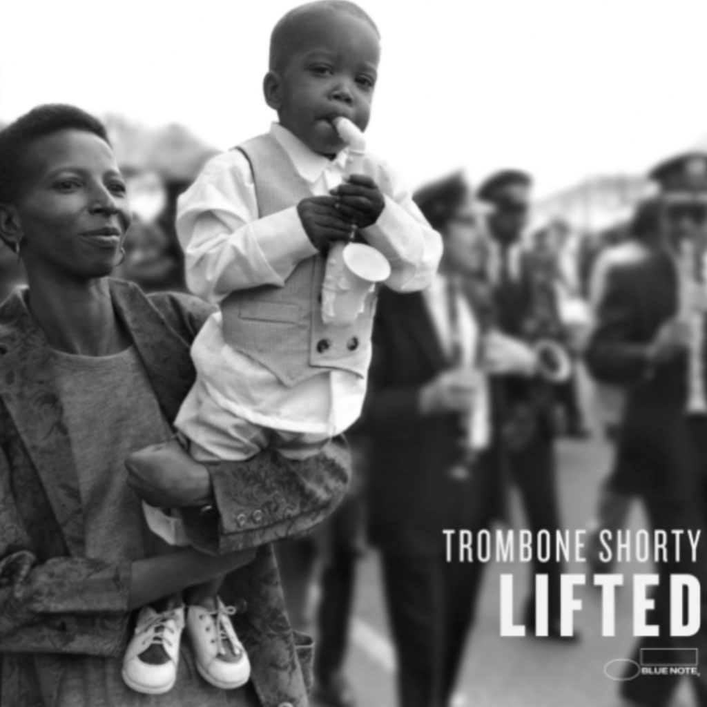 Black and white photo of a Black woman holding up a young Black child, who is playing a toy saxophone; a marching band is seen in the background