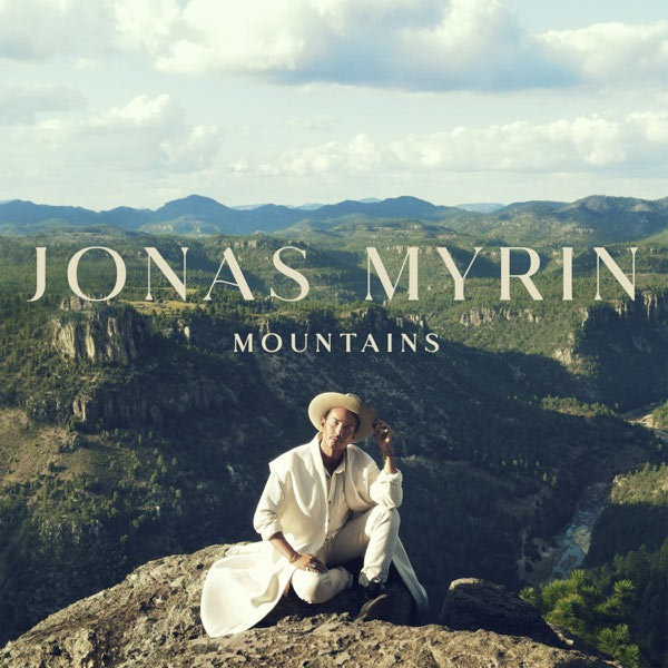 Man in white clothing and tan hat, sitting. Mountains are visible across the horizon in the background. Stylized text above the man reads, "Jonas Myrin, Mountains"