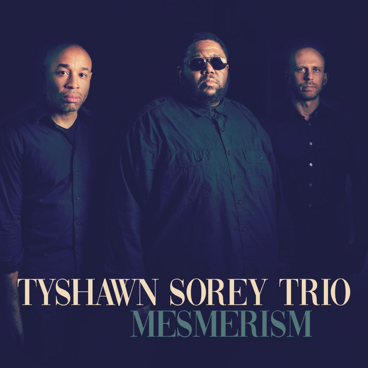 Portrait of three men standing side by side against a muted navy background. Stylized text at the bottom of the fram reads, "Tyshawn Sorey Trio, Mesmerism"